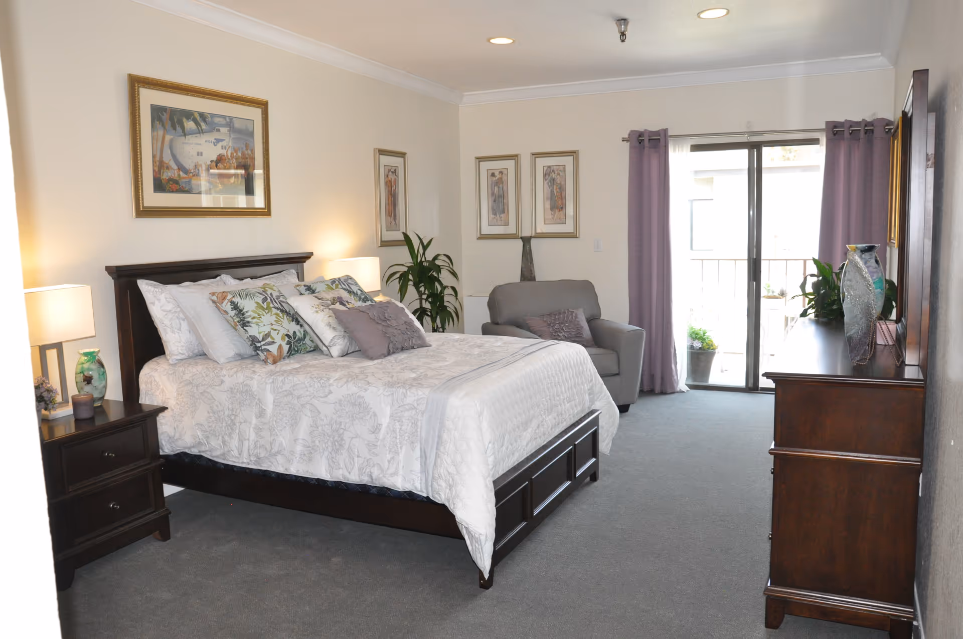 A spacious furnished bedroom with a large bed, nightstands, armchair, dresser, and sliding glass door to a balcony.