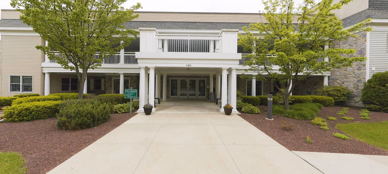 Front entrance of a building with white columns and a covered porch area, surrounded by green trees and shrubs. There is a sign near the entrance that reads 'The Commons Entrance' and a concrete walkway leading up to the doors.