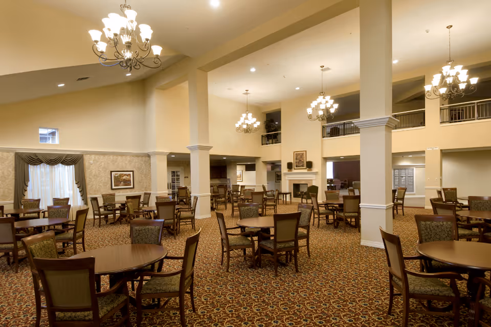 Spacious dining room with multiple round wooden tables and cushioned chairs arranged neatly on a patterned carpet. The room features high ceilings with elegant chandeliers, large columns, and a fireplace with framed artwork above it. There are windows with drapes and an upper balcony area visible.