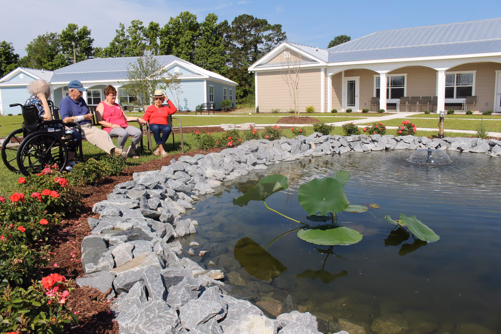Four people sit by a landscaped pond with water lilies in front of single-story cottages on a sunny day.