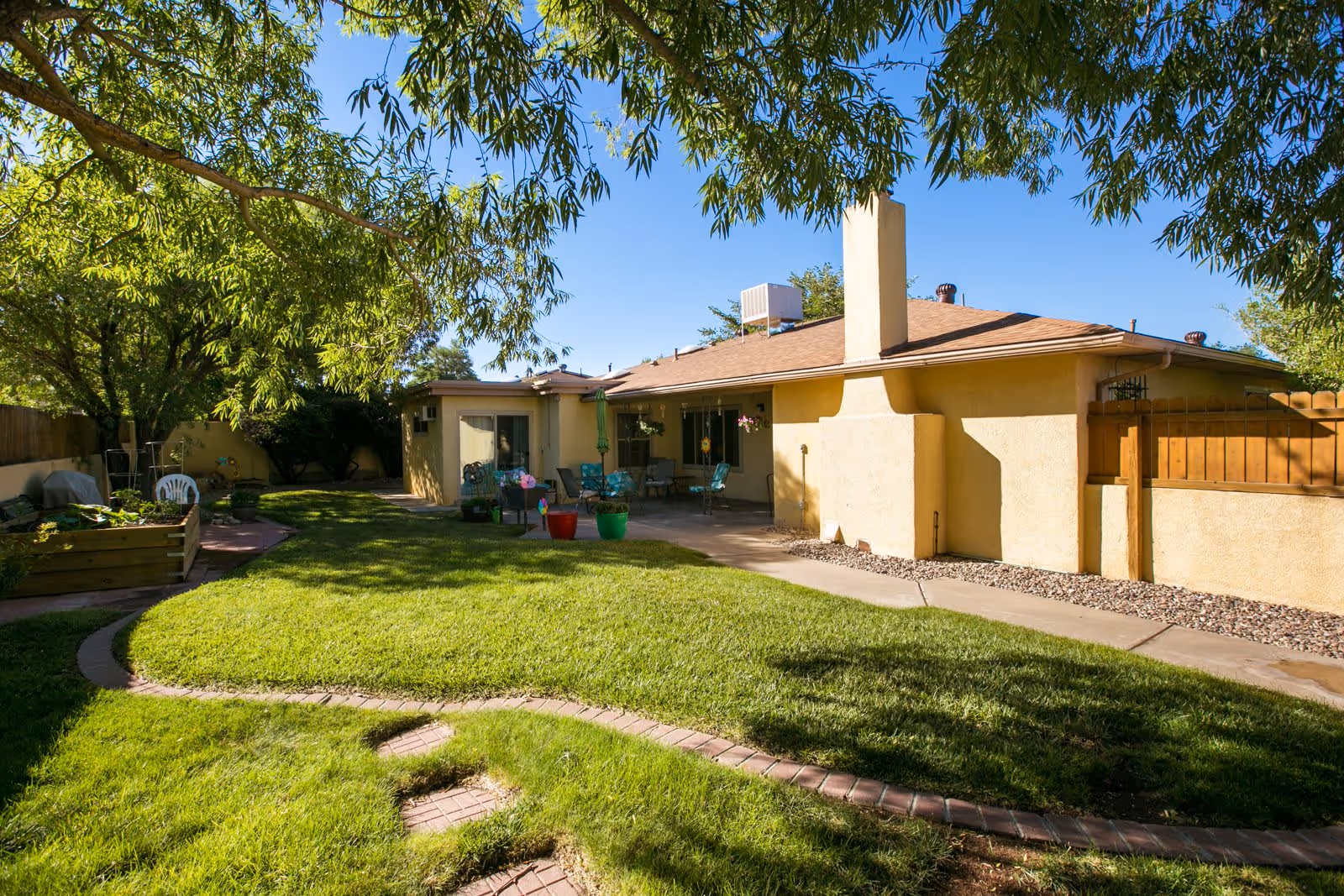 A sunny backyard area of a residential facility with a well-maintained green lawn, a curved brick pathway, and a patio with outdoor seating and potted plants. The building has a beige exterior with a chimney and is surrounded by trees and a wooden fence.