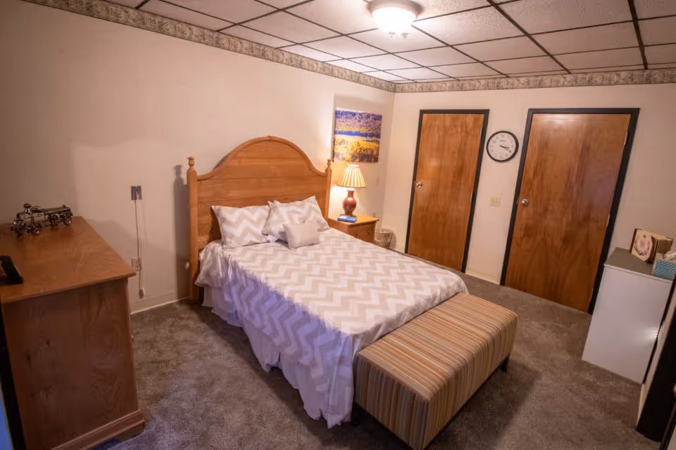A bedroom with a wooden headboard bed covered in a white and gray chevron patterned bedspread, two pillows, and a small decorative pillow. There is a striped bench at the foot of the bed, a wooden dresser on the left, a nightstand with a lamp and a picture on the wall above it, two wooden doors, a clock on the wall, and a small white cabinet with decorative items on the right. The room has beige walls, carpeted floor, and a ceiling light fixture.