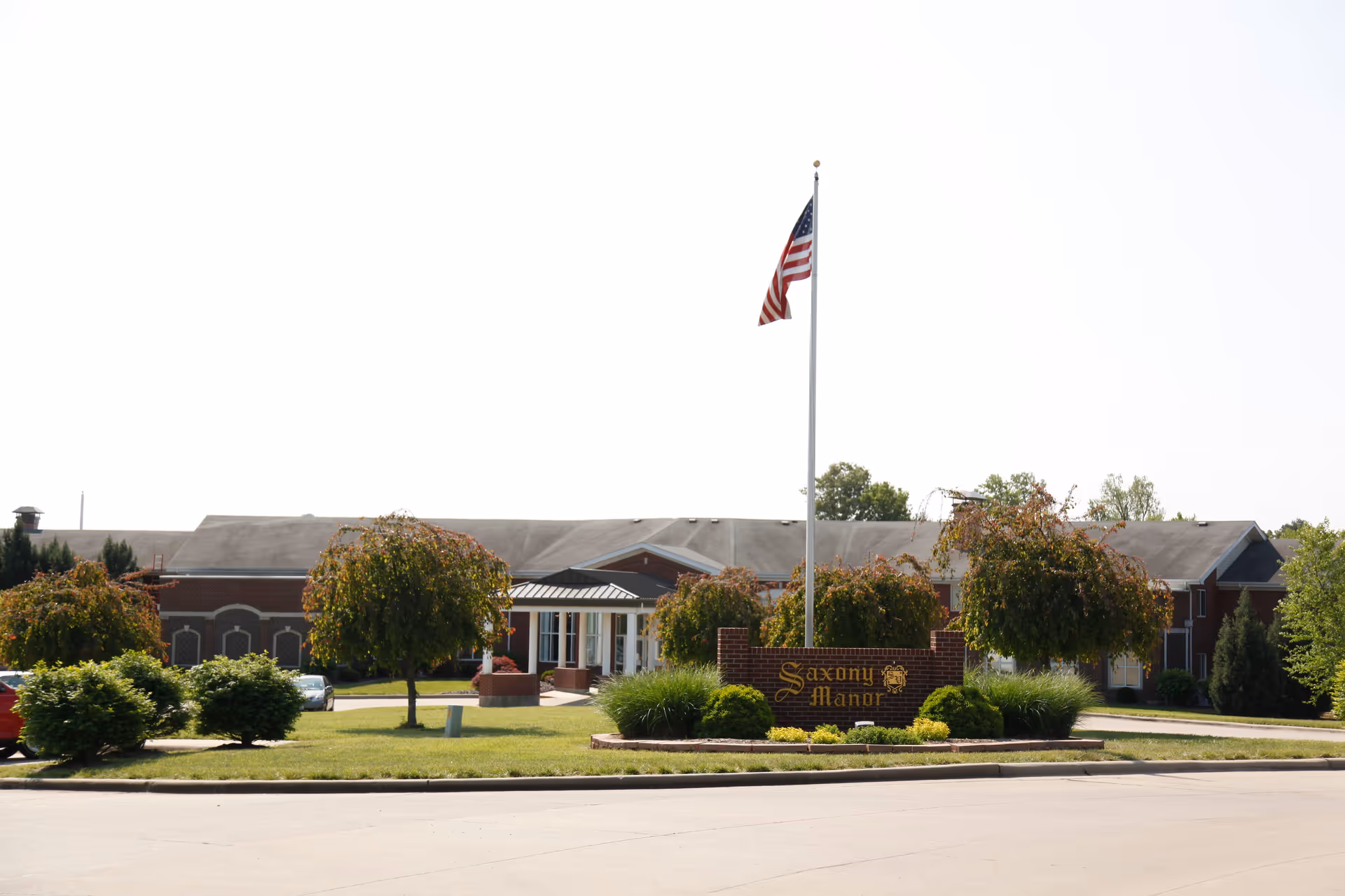 Exterior view of Saxony Manor, a brick building with a large American flag on a flagpole in front. The entrance is visible with a covered walkway, surrounded by green bushes and trees.