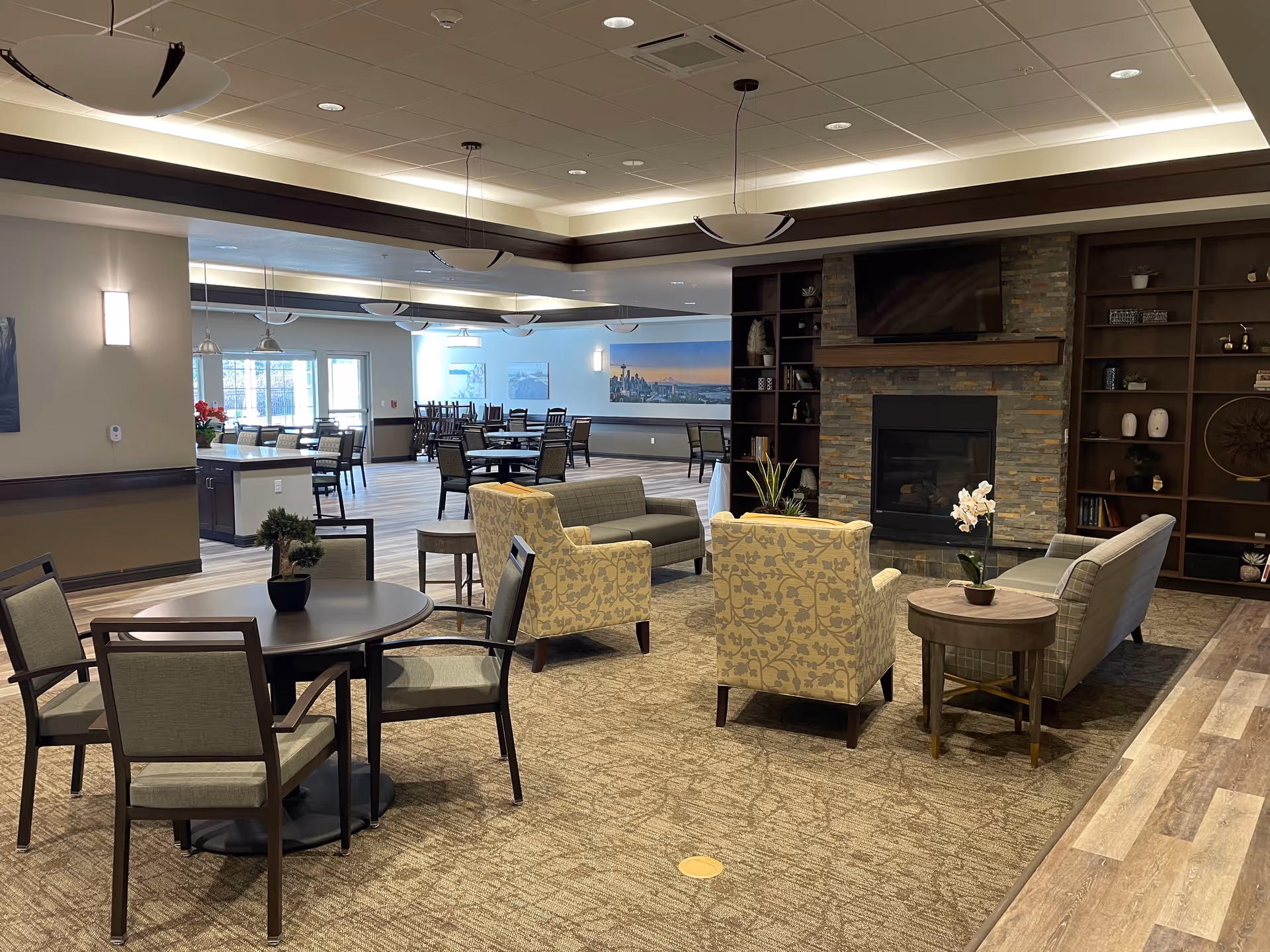A spacious and well-lit common area in a senior living facility featuring a seating arrangement with two patterned armchairs, a gray sofa, a round wooden side table with a small potted orchid, and a round table with four chairs and a small plant. In the background, there are multiple dining tables and chairs, large windows letting in natural light, and a stone fireplace with a mounted TV above it. Shelves with decorative items flank the fireplace.