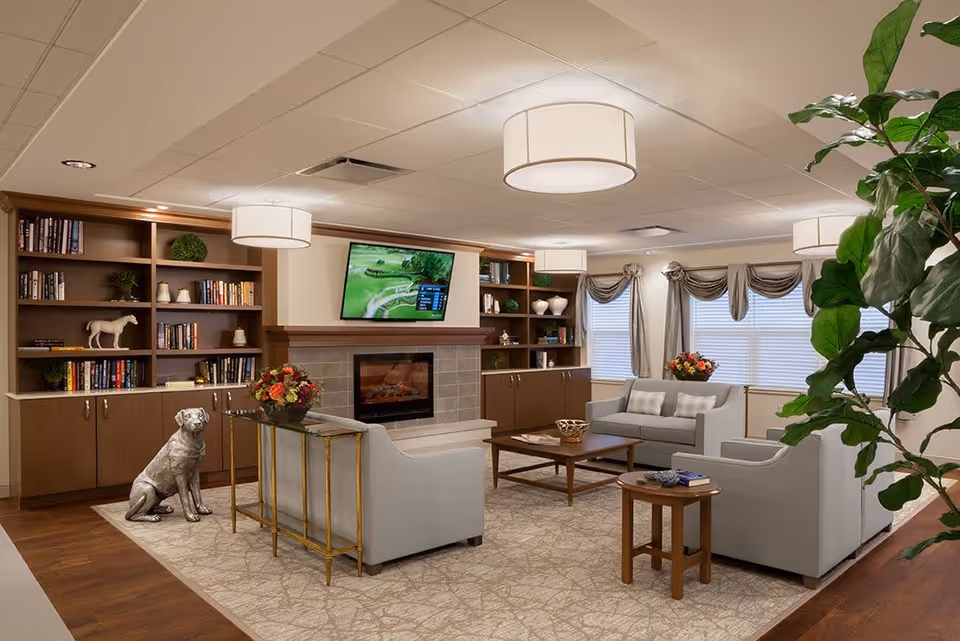 A cozy living room area with light gray sofas and armchairs arranged around a wooden coffee table and side table. There is a fireplace with a mounted flat-screen TV above it, built-in wooden bookshelves filled with books and decorative items, and large windows with draped curtains. The room is well-lit with ceiling lights and has a large indoor plant on the right side.