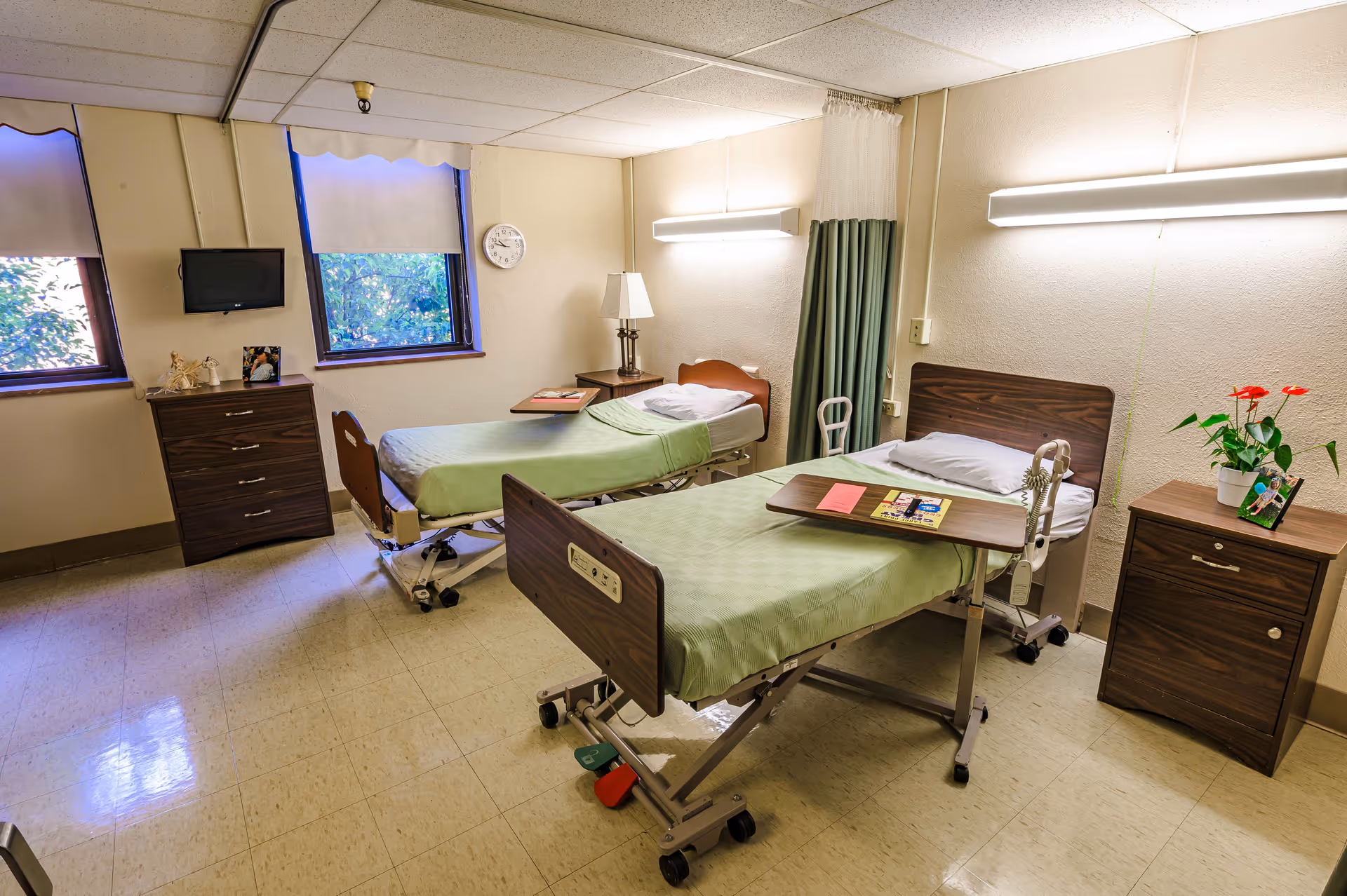 A shared patient room with two hospital beds, nightstands, a small TV, and windows.