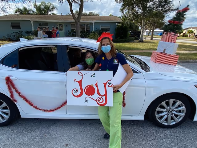 A woman wearing a red bow headband and a face mask stands next to a white car decorated with red garland and presents on the roof. She holds a sign that says 'Joy' in red letters. Another person wearing a green face mask is sitting inside the car, visible through the open window. The scene is outdoors in a residential neighborhood with houses and trees in the background.
