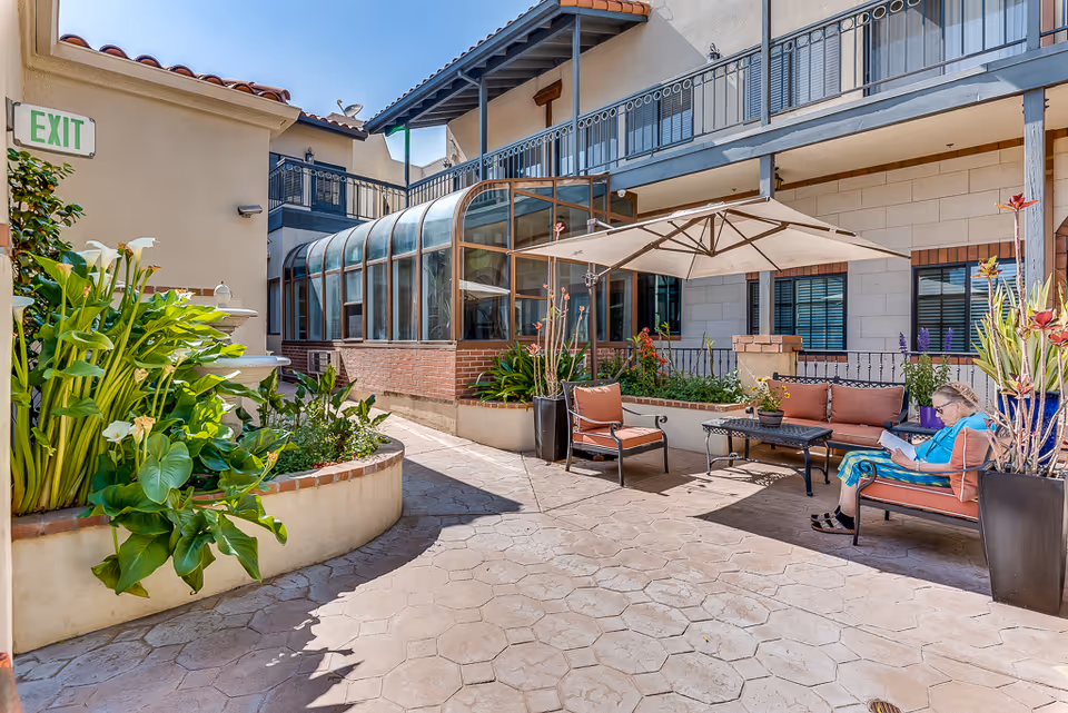 Outdoor courtyard area at The Villas by Astoria Park featuring a paved walkway, lush green plants, a seating area with cushioned chairs and a sofa under a large umbrella, and a woman sitting on one of the chairs reading a book. The building has a balcony with railings and large windows.