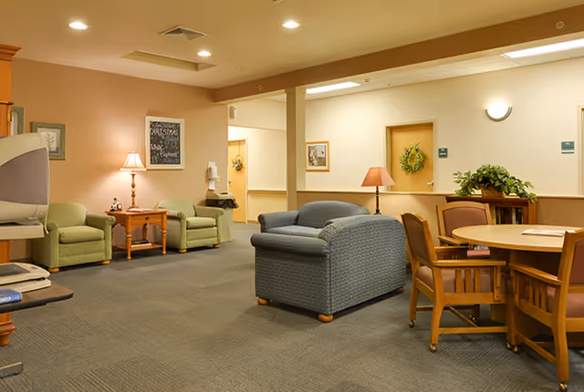 A cozy common area in a senior living facility with several upholstered chairs and a loveseat arranged around wooden tables. The walls are painted in warm tones with framed artwork and wreath decorations. There are lamps providing soft lighting and a carpeted floor.