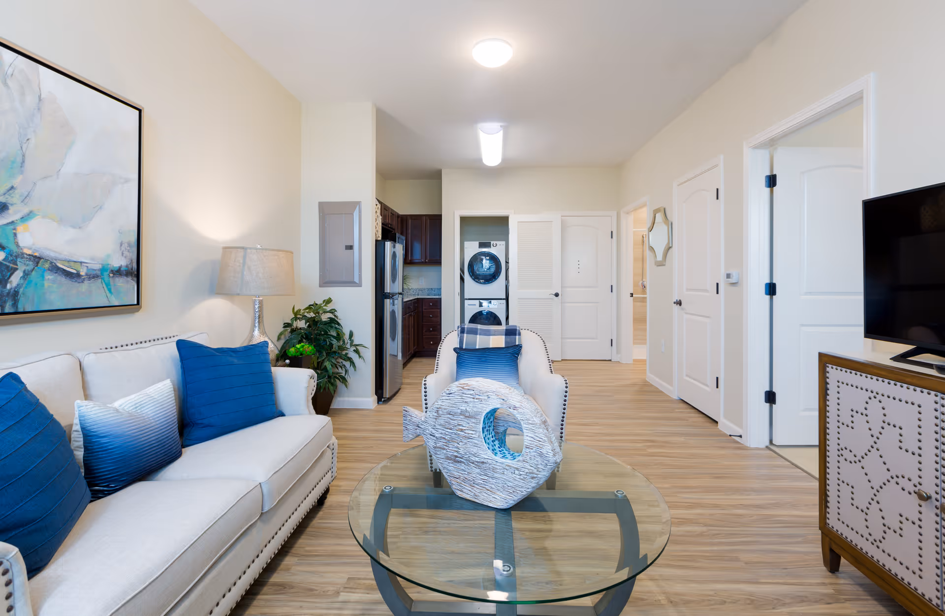 A bright and modern living room in a senior living facility featuring a beige sofa with blue and white pillows, a glass coffee table with a decorative sculpture, a matching armchair, a flat-screen TV on a wooden cabinet, and a view into a kitchen area with stainless steel appliances and a stacked washer and dryer unit.