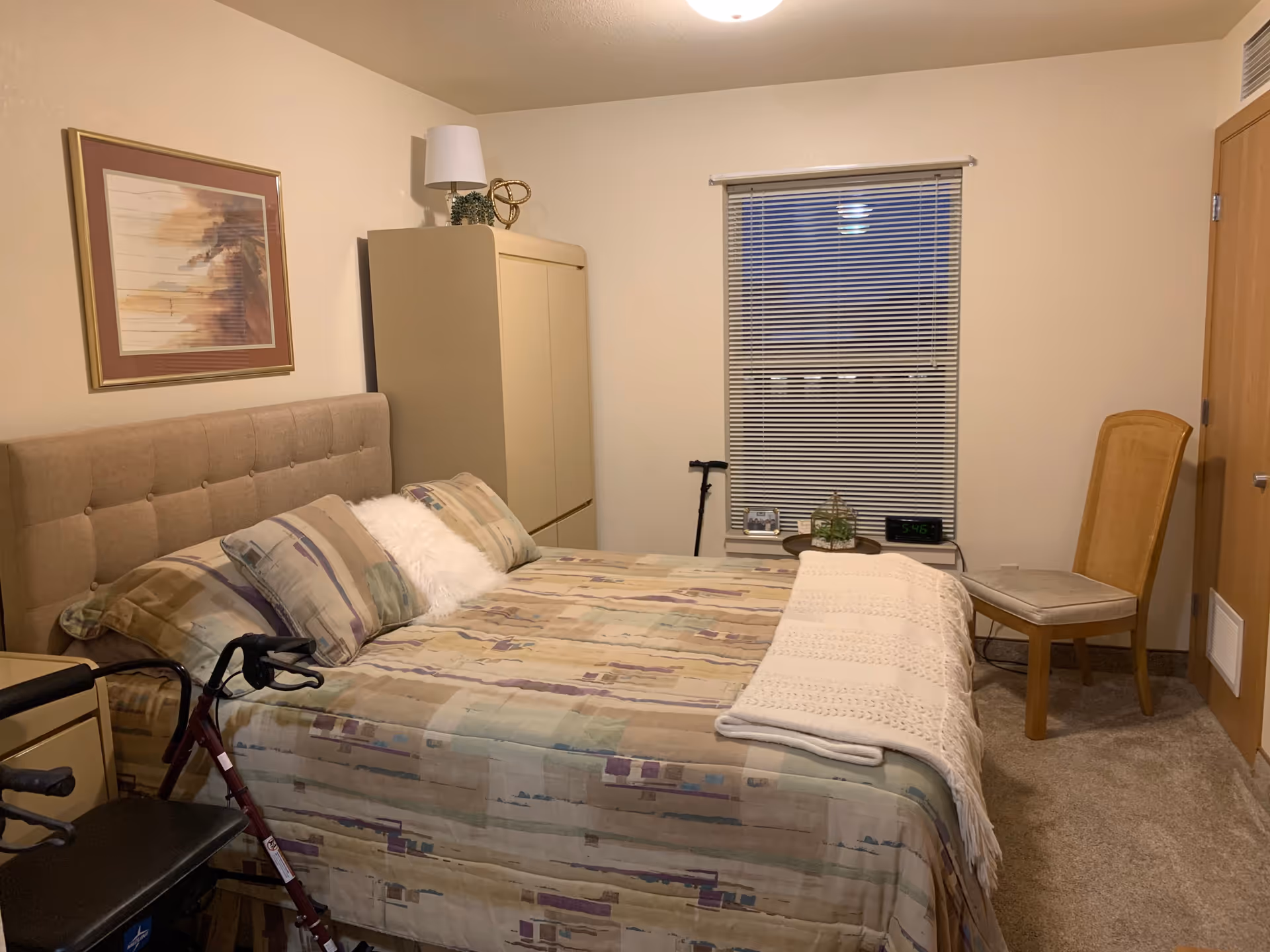 A bedroom with a large bed covered in a patterned bedspread and pillows. There is a beige headboard, a beige wardrobe with a lamp and decorative item on top, a window with closed blinds, a wooden chair, and a walking aid near the bed. The room has beige walls and carpeted floor.