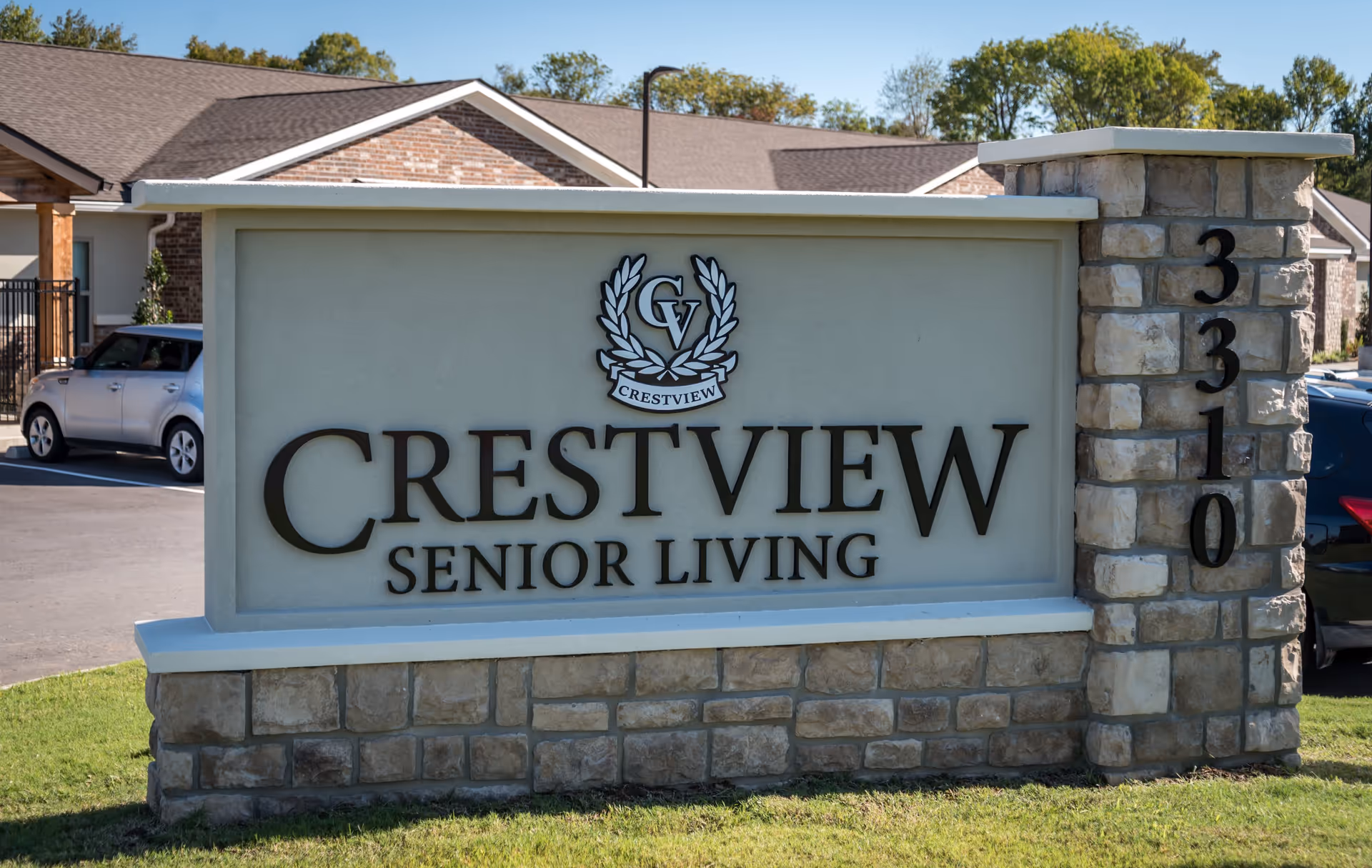 Stone and concrete sign for Crestview Senior Living with the address number 3310 on a stone pillar, set in front of a parking lot and buildings with trees in the background.