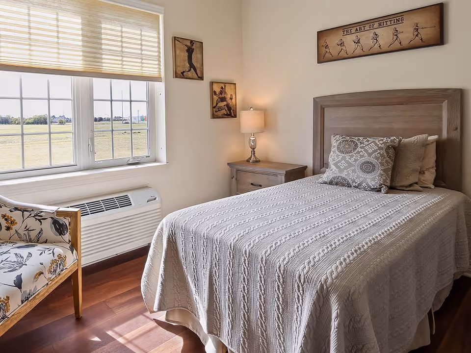 A cozy bedroom with a wooden bed frame and a neatly made bed covered with a textured beige blanket and decorative pillows. Next to the bed is a wooden nightstand with a lamp. The room has a large window with blinds partially drawn, letting in natural light. There is a floral-patterned armchair near the window. The walls are decorated with three framed pictures, including one above the bed titled 'The Art of Hitting' showing a sequence of a baseball player swinging a bat.
