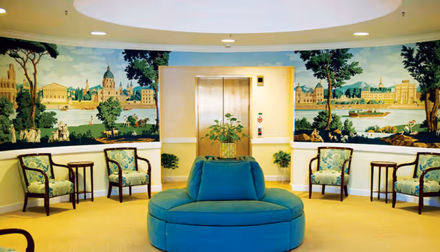 A seating area in a senior living facility with a circular blue cushioned bench in the center, surrounded by four wooden chairs with floral upholstery and small side tables. The back wall features an elevator door framed by a mural depicting a scenic riverside cityscape with trees, buildings, and people.