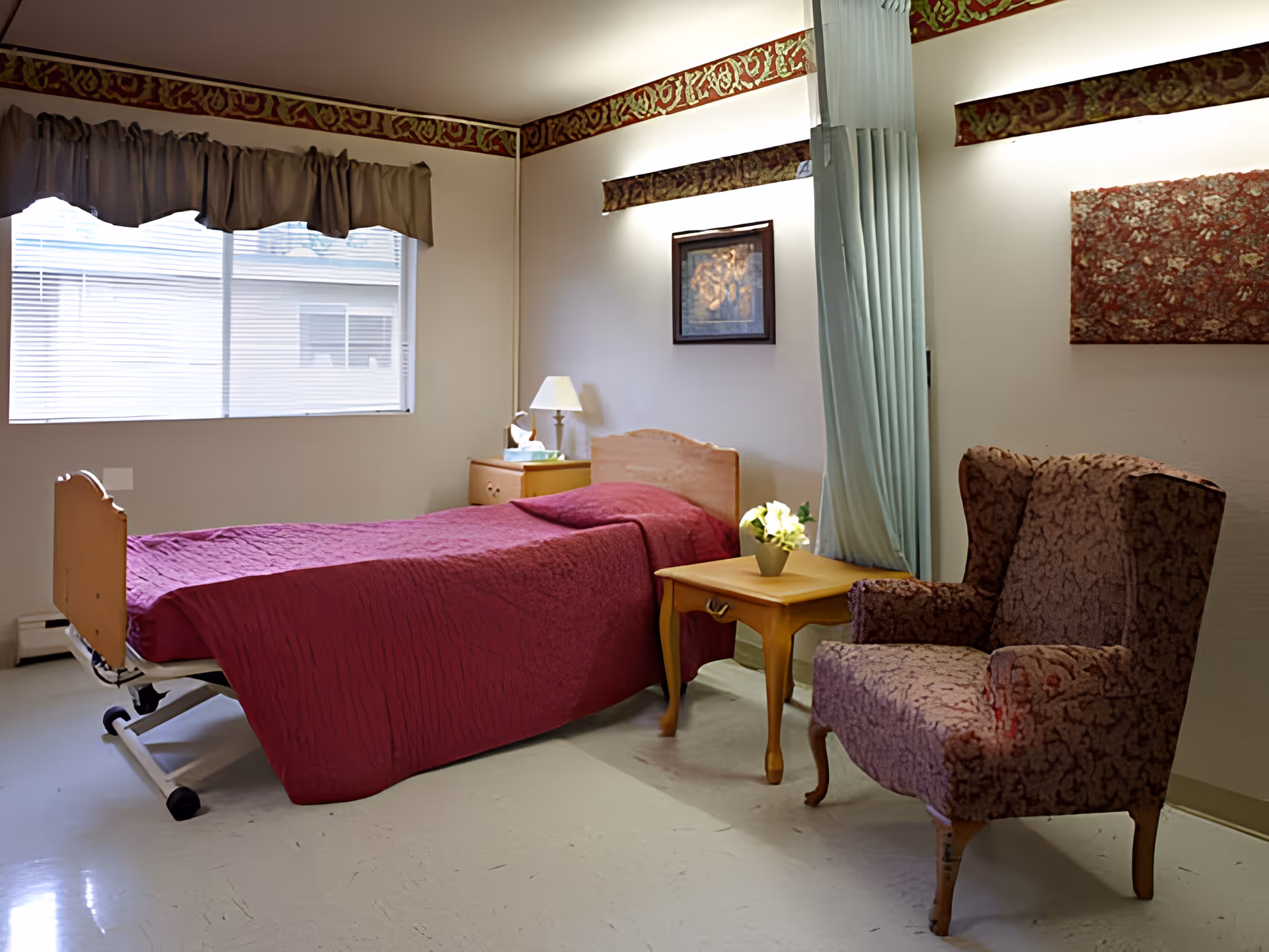 A simple, clean bedroom in a healthcare facility featuring a single bed with a red bedspread, a wooden headboard, a bedside table with a lamp and tissues, a small wooden table with a potted plant, and a patterned armchair. The room has a large window with blinds and a curtain, decorative wall borders near the ceiling, and framed artwork on the walls.