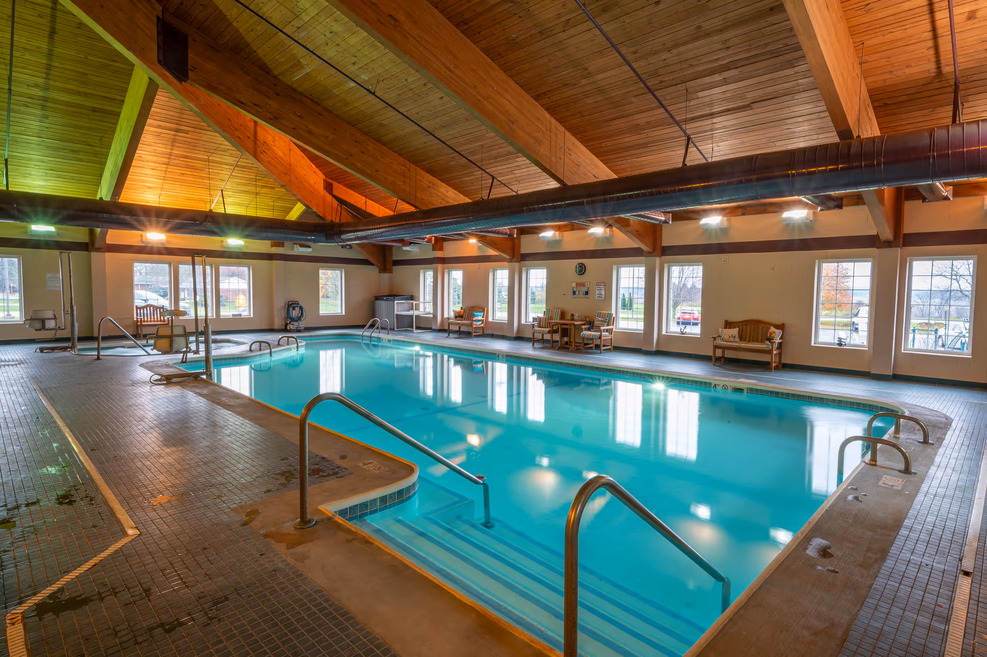 Indoor swimming pool with clear blue water, surrounded by tiled flooring and wooden benches along the walls. The ceiling features exposed wooden beams and large windows let in natural light.