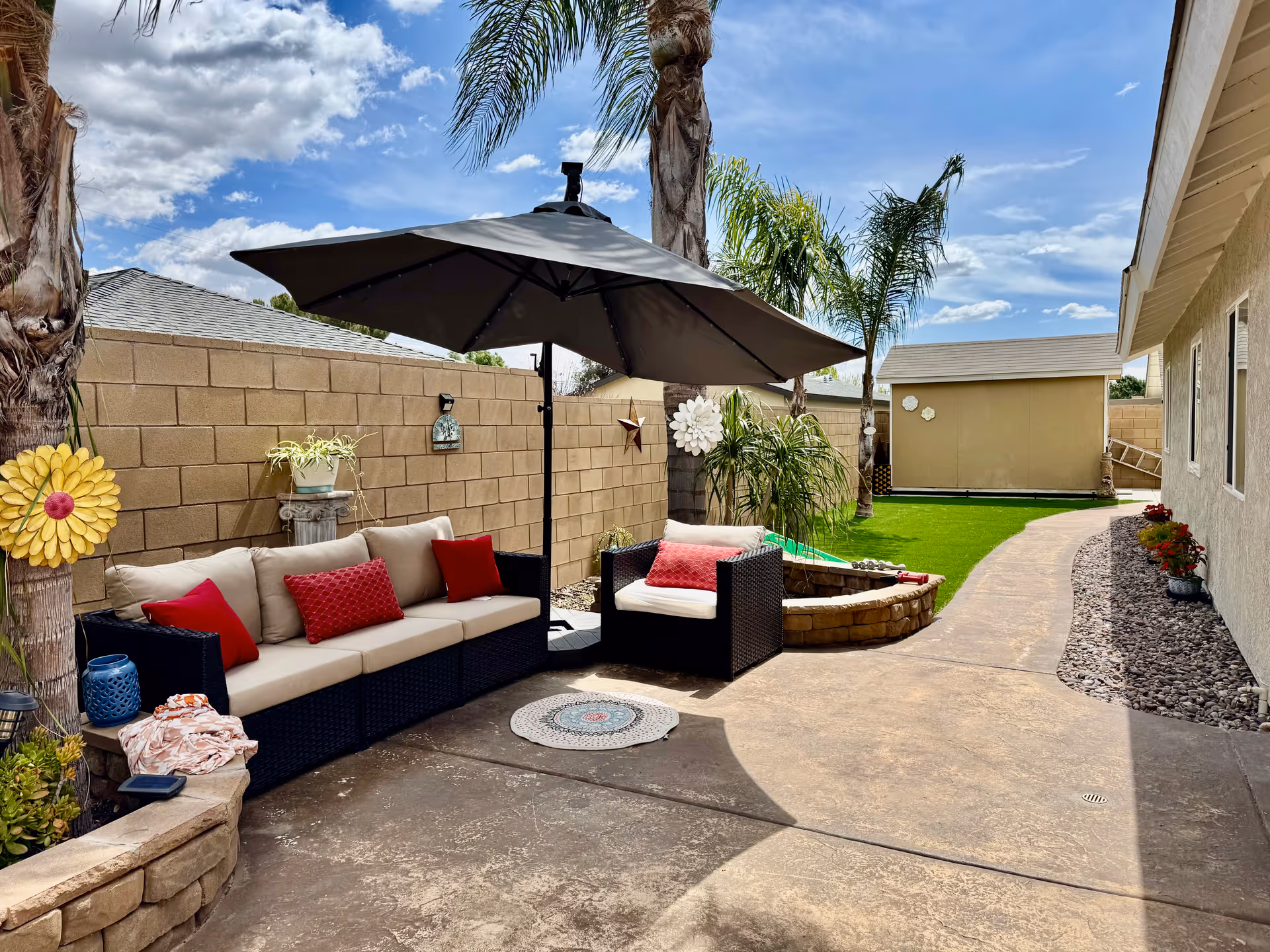 Outdoor patio area with a beige cushioned sofa and armchair with red pillows under a large black umbrella. The patio is surrounded by a beige brick wall, palm trees, and decorative wall hangings. There is a curved concrete pathway leading to a small shed in the background, with green artificial grass and some potted plants along the side of a beige building.