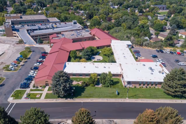 Aerial view of Gateway Transitional Care Center showing a large building complex with red and white roofs, surrounded by green lawns, trees, and parking lots. The building is situated in a suburban area with many trees and houses visible in the background.