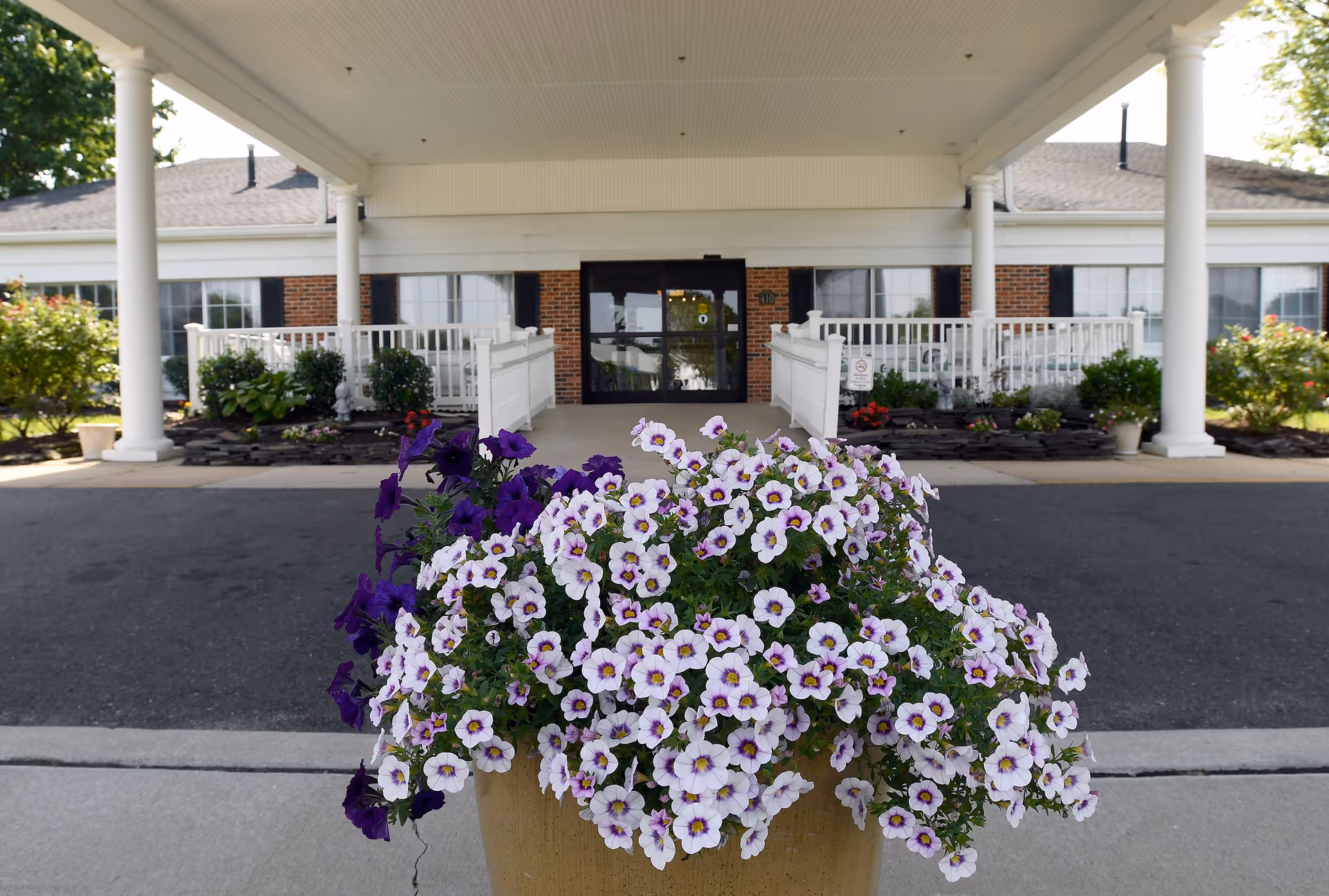 Entrance of a senior living facility with a covered driveway, white columns, and a large planter filled with purple and white flowers in the foreground. The building has brick walls and multiple windows with black shutters.