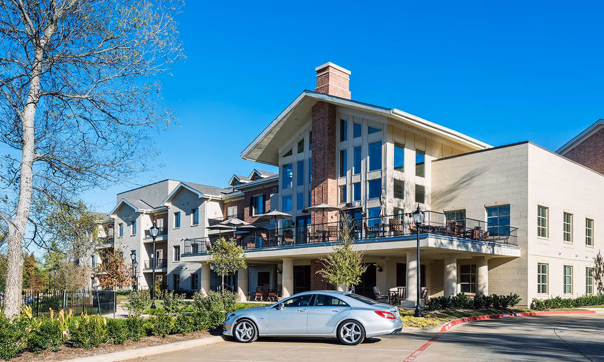 Exterior view of a senior living facility building with a modern design featuring large windows, a balcony with patio umbrellas and seating, surrounded by trees and landscaping under a clear blue sky. A silver car is parked in front of the building.
