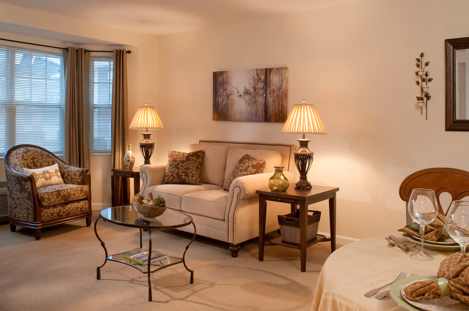 A cozy living room with a beige sofa adorned with patterned cushions, a matching armchair, two side tables each with a lamp, a glass coffee table with decorative items, and a dining table set with plates, napkins, and wine glasses. The room has beige walls, a window with blinds and curtains, and wall decorations including a painting and a mirror.