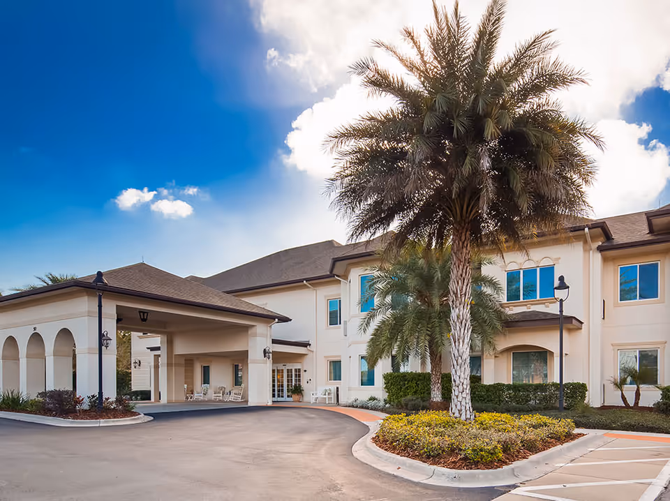 Front entrance of a two-story beige senior living building with a covered porte-cochere, palm trees, and a curved driveway under a blue sky.