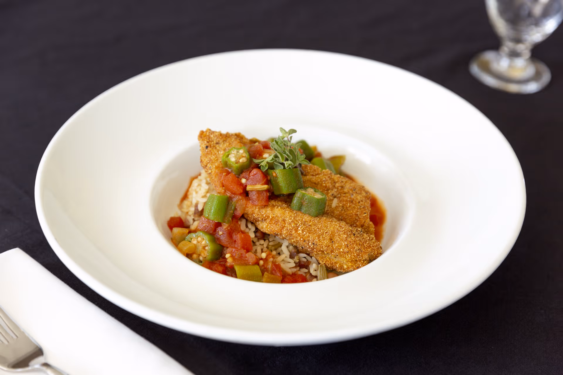 A white bowl containing a serving of rice topped with fried fish fillets, okra slices, diced tomatoes, and garnished with a small sprig of herbs, placed on a black tablecloth with a fork and napkin nearby and a glass of water in the background.