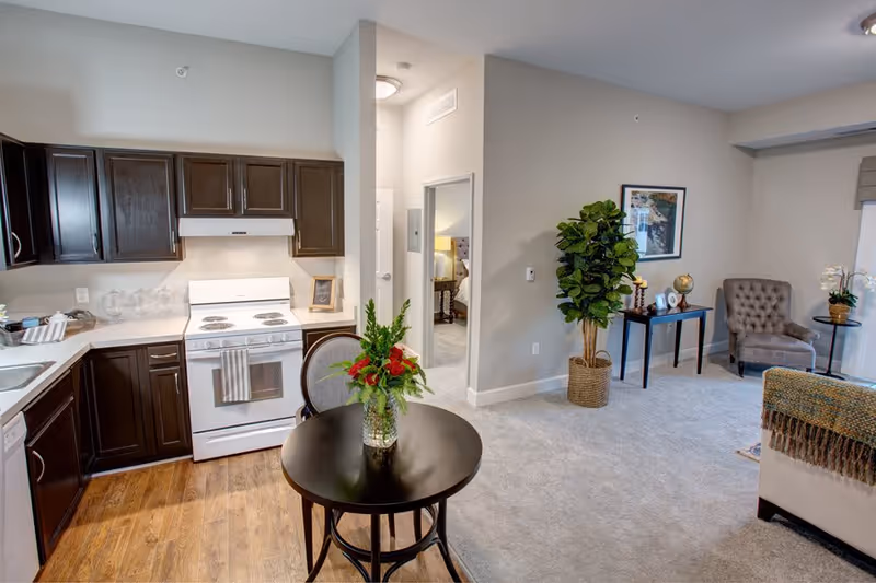 Interior view of a senior living facility apartment showing a kitchen area with dark wood cabinets, a white stove, and a countertop. Adjacent to the kitchen is a small round table with a vase of flowers. The living area has carpeted flooring, a potted plant, a framed picture on the wall, a small table with decorative items, and a gray upholstered armchair near a window with blinds.