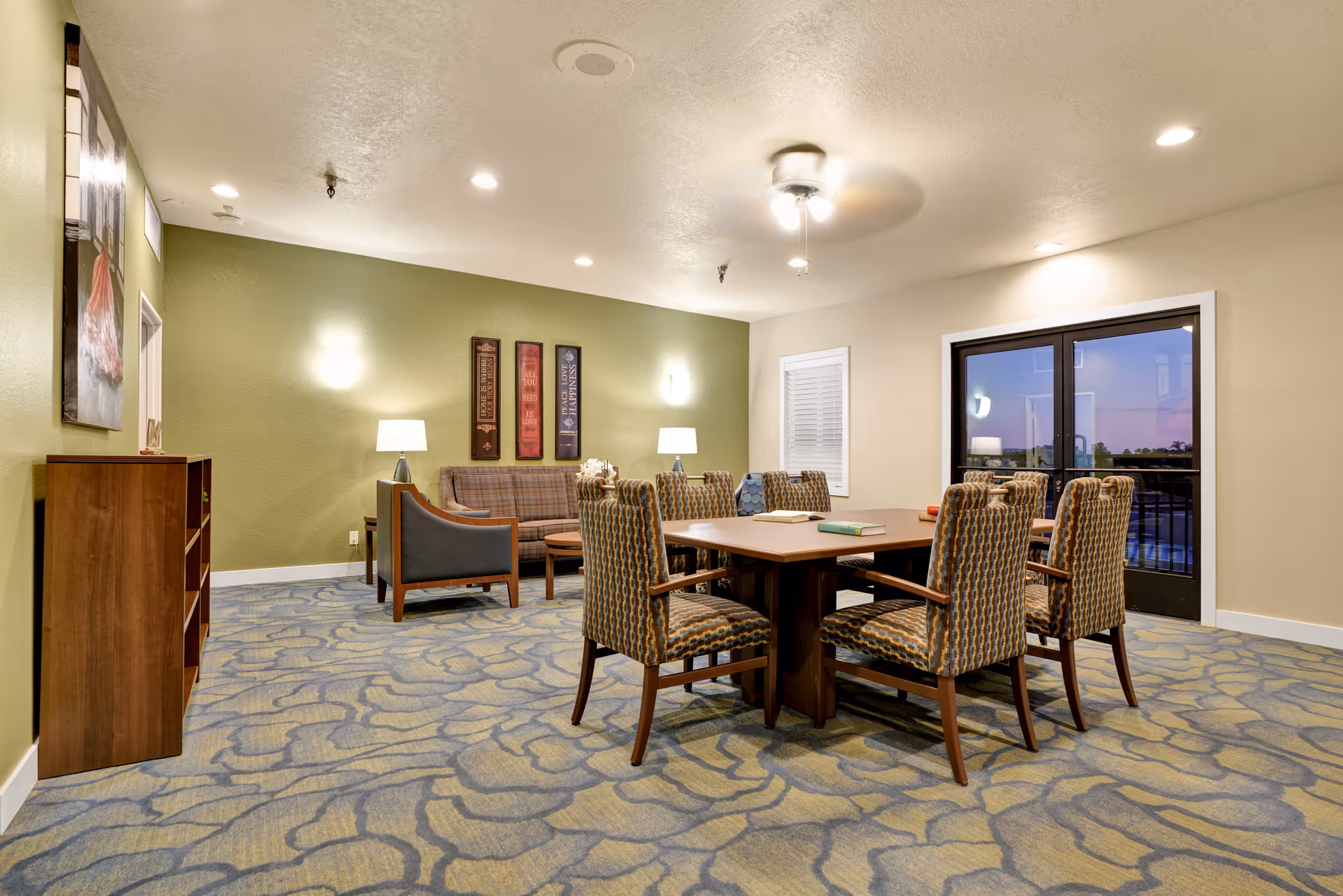 A well-lit common room with a large wooden table surrounded by six patterned chairs. In the background, there is a seating area with a plaid sofa, two armchairs, two table lamps, and three framed wall decorations on a green accent wall. The room has a patterned carpet, beige walls, a ceiling fan with lights, and a glass door leading outside.