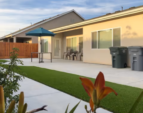 Outdoor patio area at Genialcare senior living facility with seating, umbrella, and trash bins.