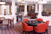 A spacious, well-lit common area with a patterned carpet, featuring a round poker table with red cushioned chairs in the foreground and a rectangular dining table with chairs in the background. The room has white columns, large windows with valances, and decorative plants, creating a comfortable and inviting atmosphere.