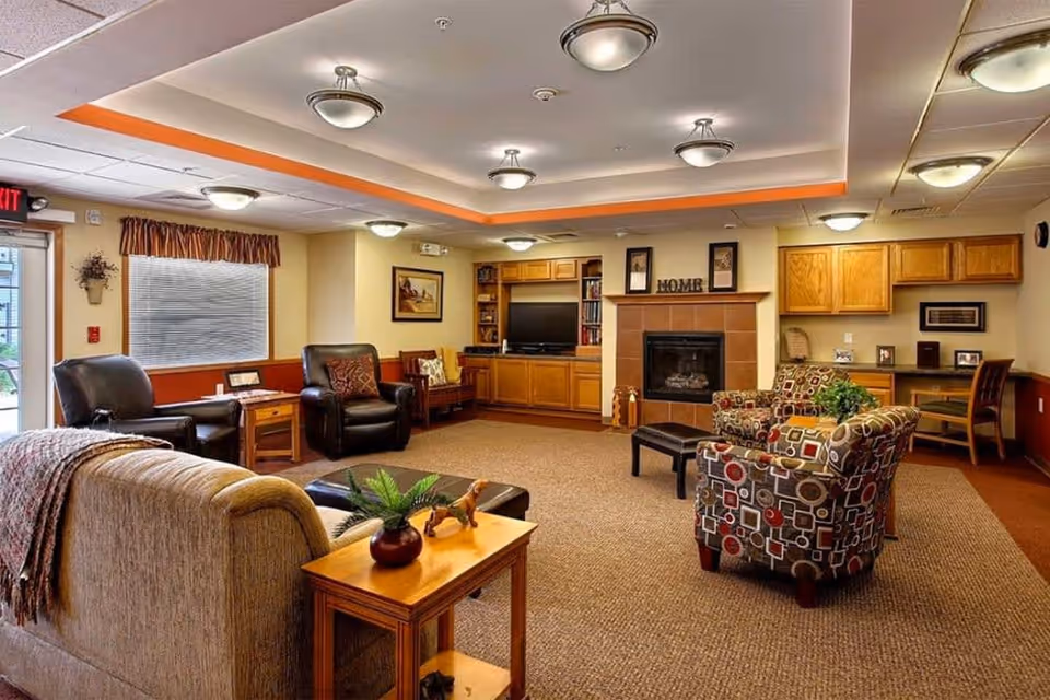 A cozy senior living facility common area with comfortable seating including a beige sofa, two patterned armchairs, and two black leather chairs. The room features a fireplace with a mantle decorated with the word 'HOME', built-in wooden cabinets, a flat-screen TV, a small desk area, and warm lighting from ceiling fixtures. There is a window with blinds and a door with an exit sign above it.
