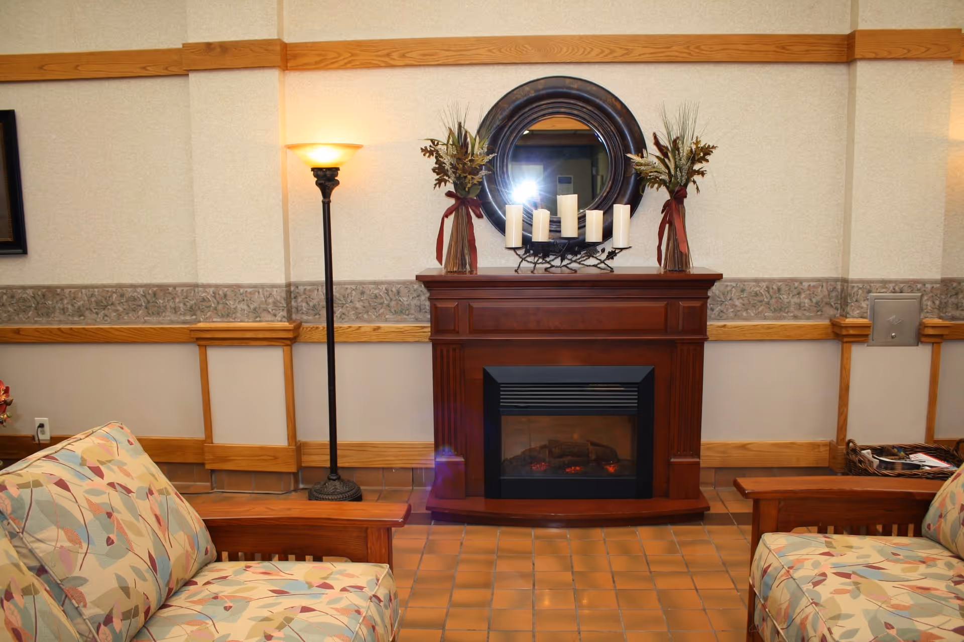 Cozy sitting area with patterned armchairs, a floor lamp, and a wooden electric fireplace topped with candles and a round mirror.