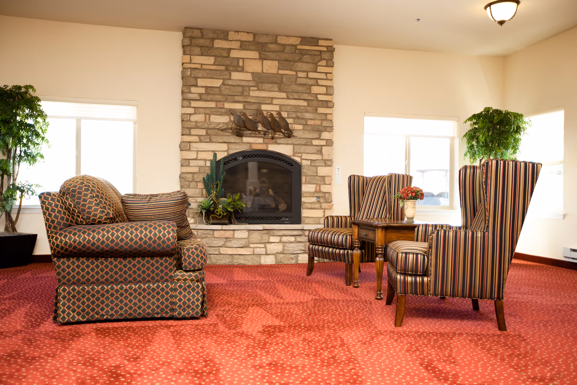 Cozy lounge area with upholstered armchairs arranged around a stone fireplace on red carpeting.