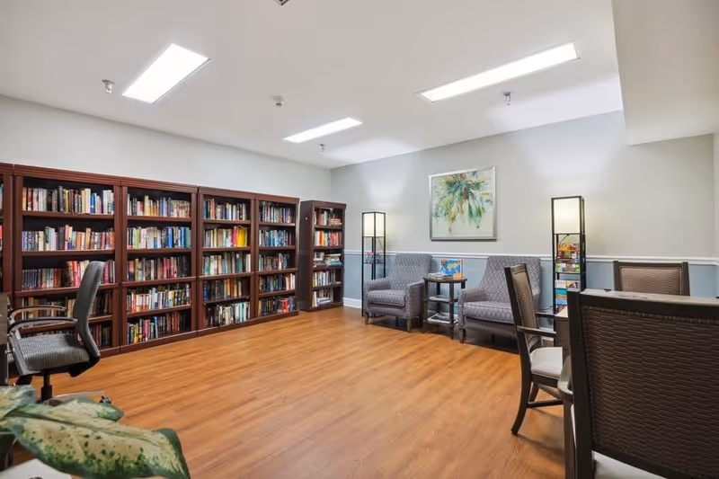 A cozy reading room with wooden bookshelves filled with books along one wall, two cushioned armchairs with a small round table between them, two tall floor lamps, and several chairs around a table. The room has wooden flooring and a framed abstract painting on the wall.