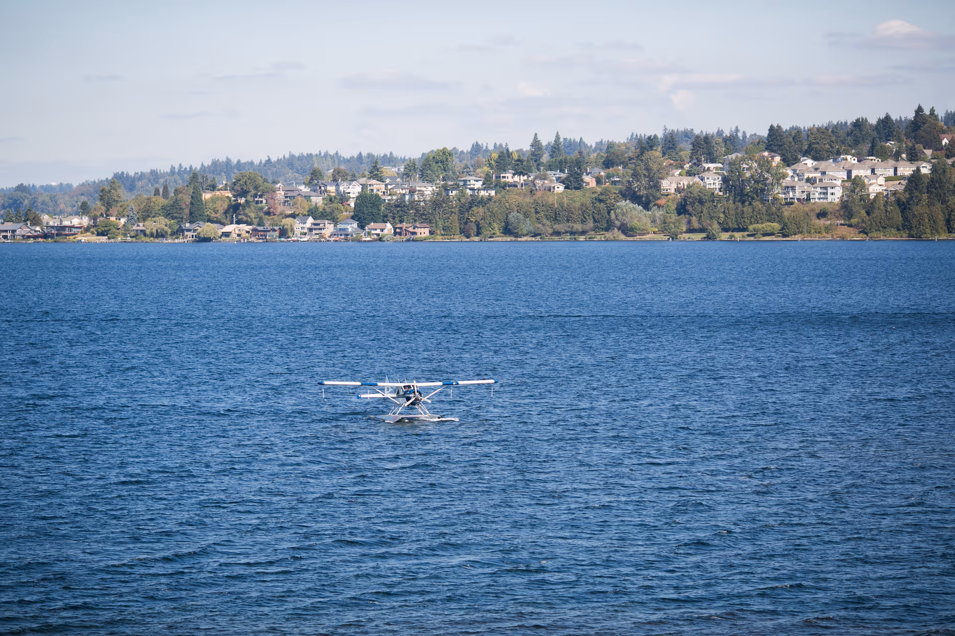 A small seaplane floating on a large body of water with a shoreline in the background featuring houses and trees under a partly cloudy sky.