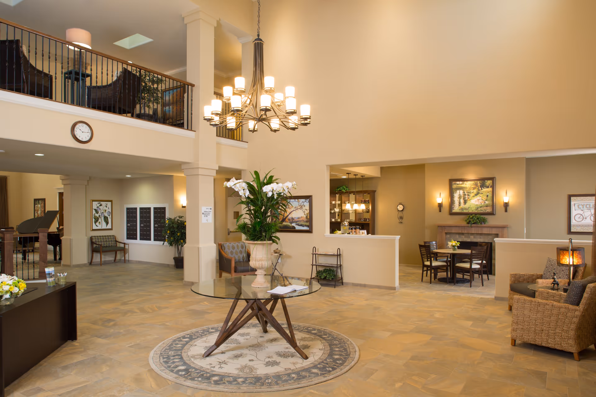Spacious and well-lit senior living facility common area with high ceilings, a large chandelier, a round glass table with a floral arrangement in the center, comfortable seating including chairs and a sofa, a fireplace with artwork above it, and a piano in the background near a staircase with a railing.