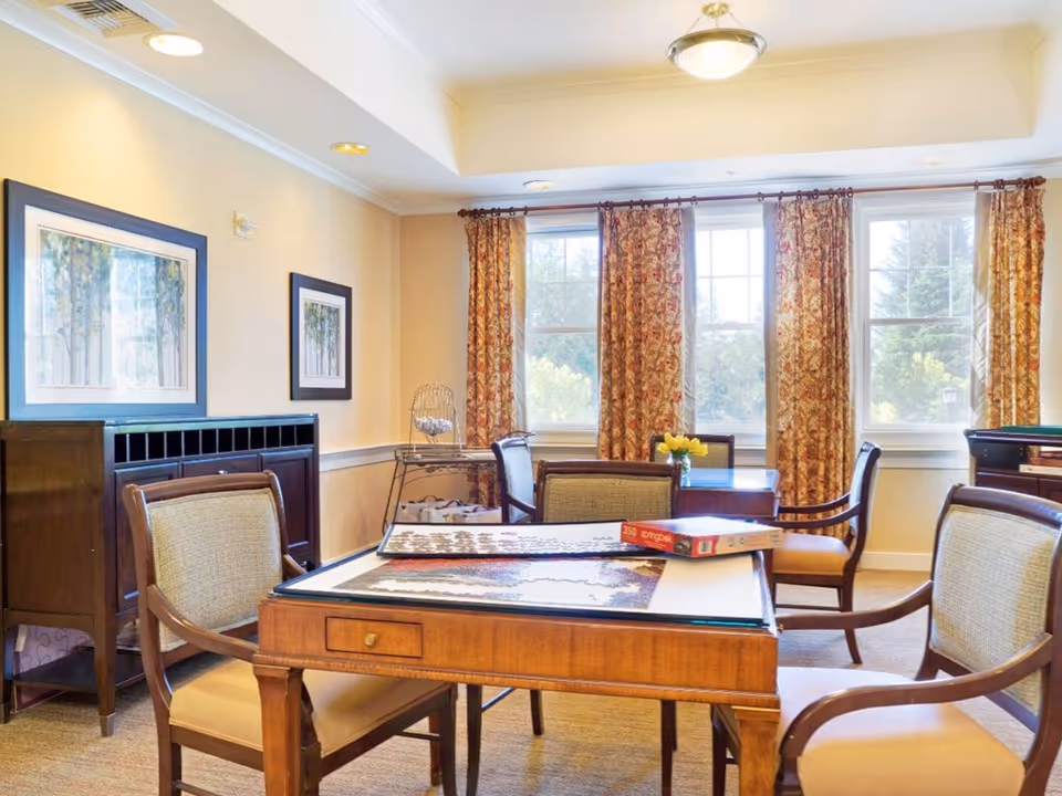 A bright and cozy common room with large windows covered by patterned curtains. The room features wooden tables and cushioned chairs arranged for social activities. On one table, there is a partially completed jigsaw puzzle and a puzzle box. The walls have framed artwork, and there is a small cabinet and a metal rack in the corner.