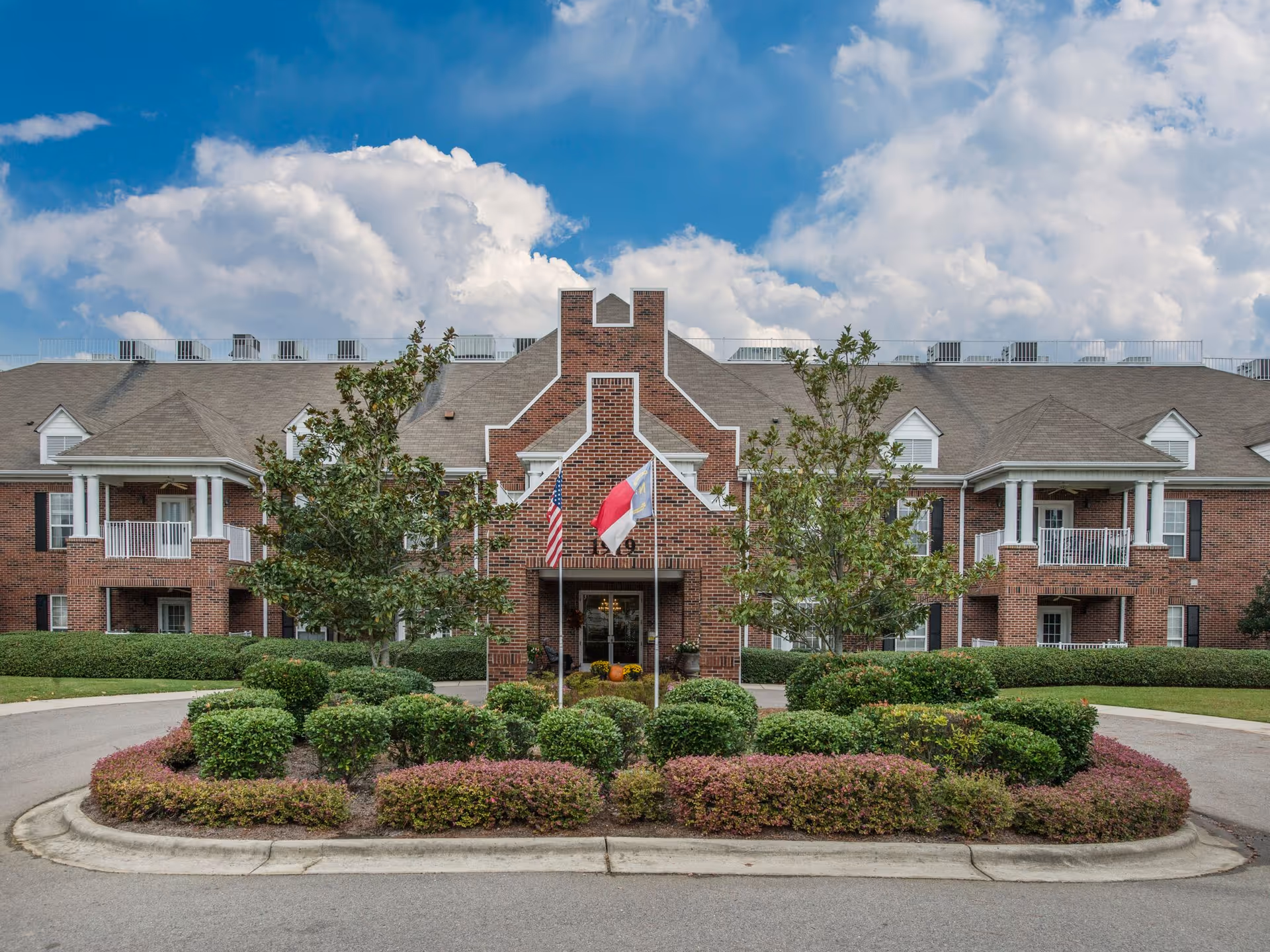 Brick senior living building front with a central entrance, flags, balconies, and landscaped roundabout under a blue sky.