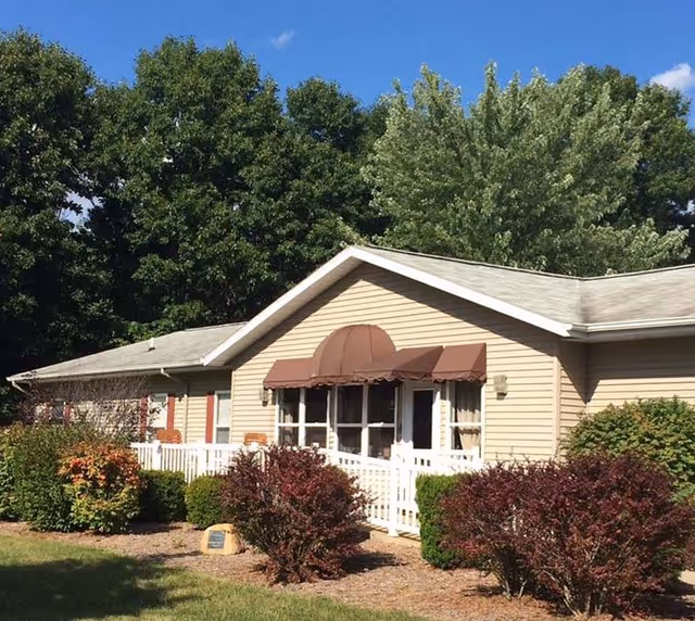 Single-story beige building with a brown awning over the entrance, surrounded by green and reddish bushes and trees under a clear blue sky.