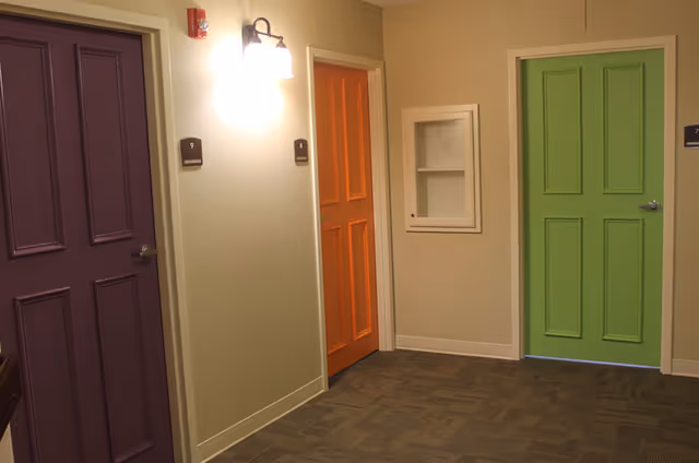 Interior hallway with three closed doors painted in different colors: purple, orange, and green. The walls are beige, and there is a wall-mounted light fixture above the orange door. The floor is carpeted with a patterned design.