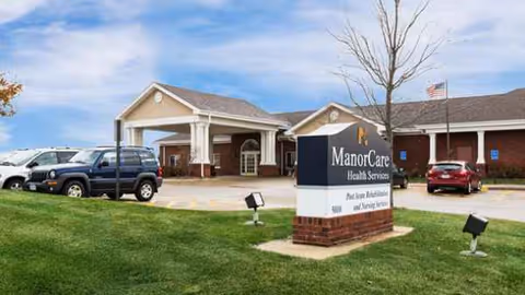 Exterior of ManorCare Health Services facility with a sign and parked vehicles in front.