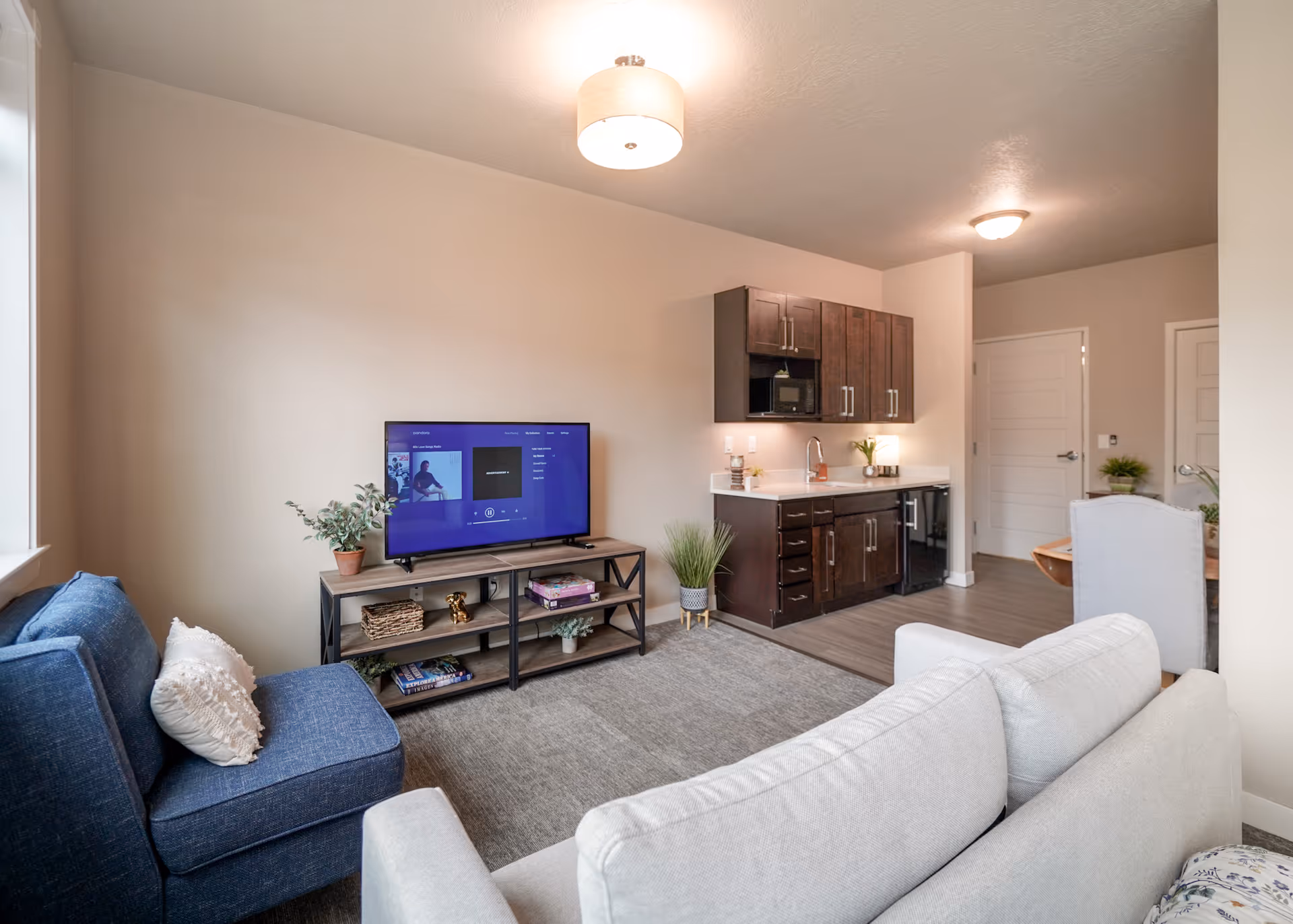Open living room with a sofa and armchair facing a TV on a stand, and a small kitchenette with dark cabinets in the background.