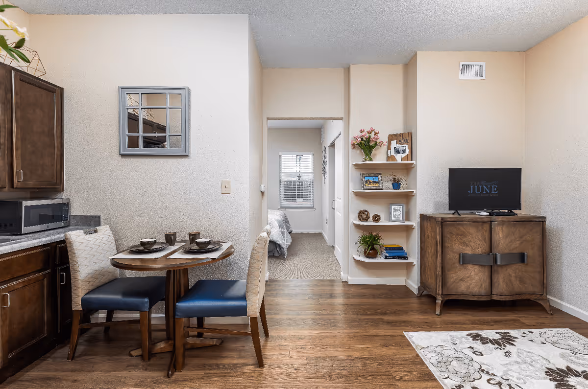 Interior view of a senior living facility showing a small dining area with a round wooden table set for two, adjacent to a kitchen counter with a microwave. The room has wooden flooring and beige walls. A hallway leads to a bedroom with a window and a bed visible. On the right side, there is a wooden cabinet with a TV on top and wall-mounted shelves with decorative items and flowers.