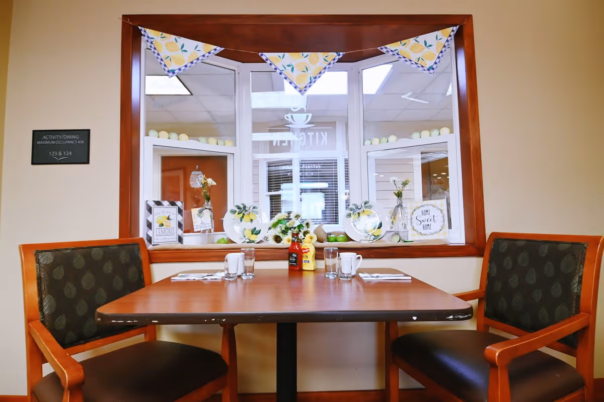 Small dining table with two chairs set by a window decorated with plates, flowers, and bunting.
