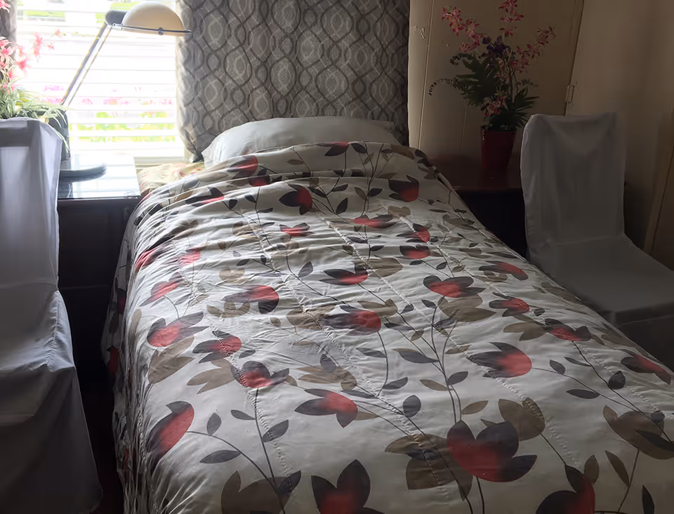 A single bed with a floral patterned comforter in shades of red, brown, and gray. The bed is positioned against a window with patterned curtains. There are two chairs covered with white fabric on either side of the bed, a small table with a lamp and a potted plant near the window, and another table with a potted plant on the right side of the bed.