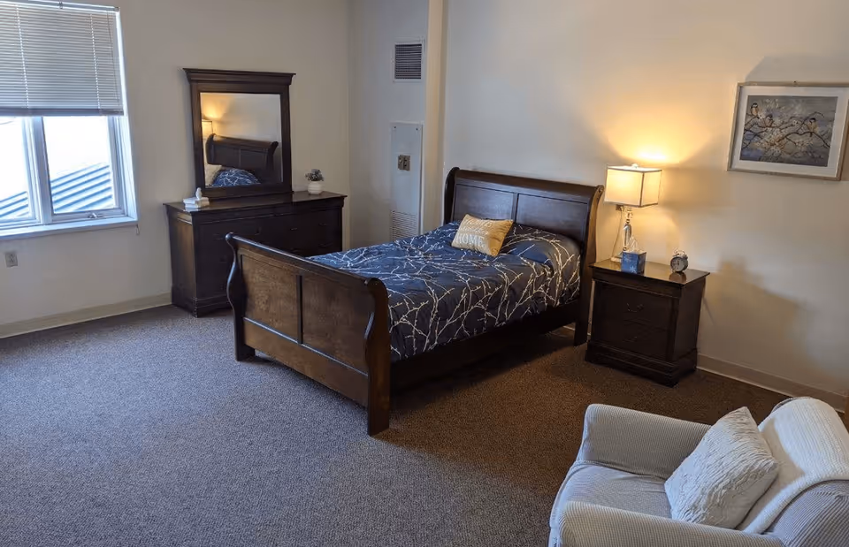 A bedroom with a wooden bed frame and blue patterned bedding, a matching wooden dresser with a mirror, a nightstand with a lamp, an alarm clock, and a tissue box. There is a window with blinds, a framed picture on the wall, and a cushioned armchair with a pillow in the corner.