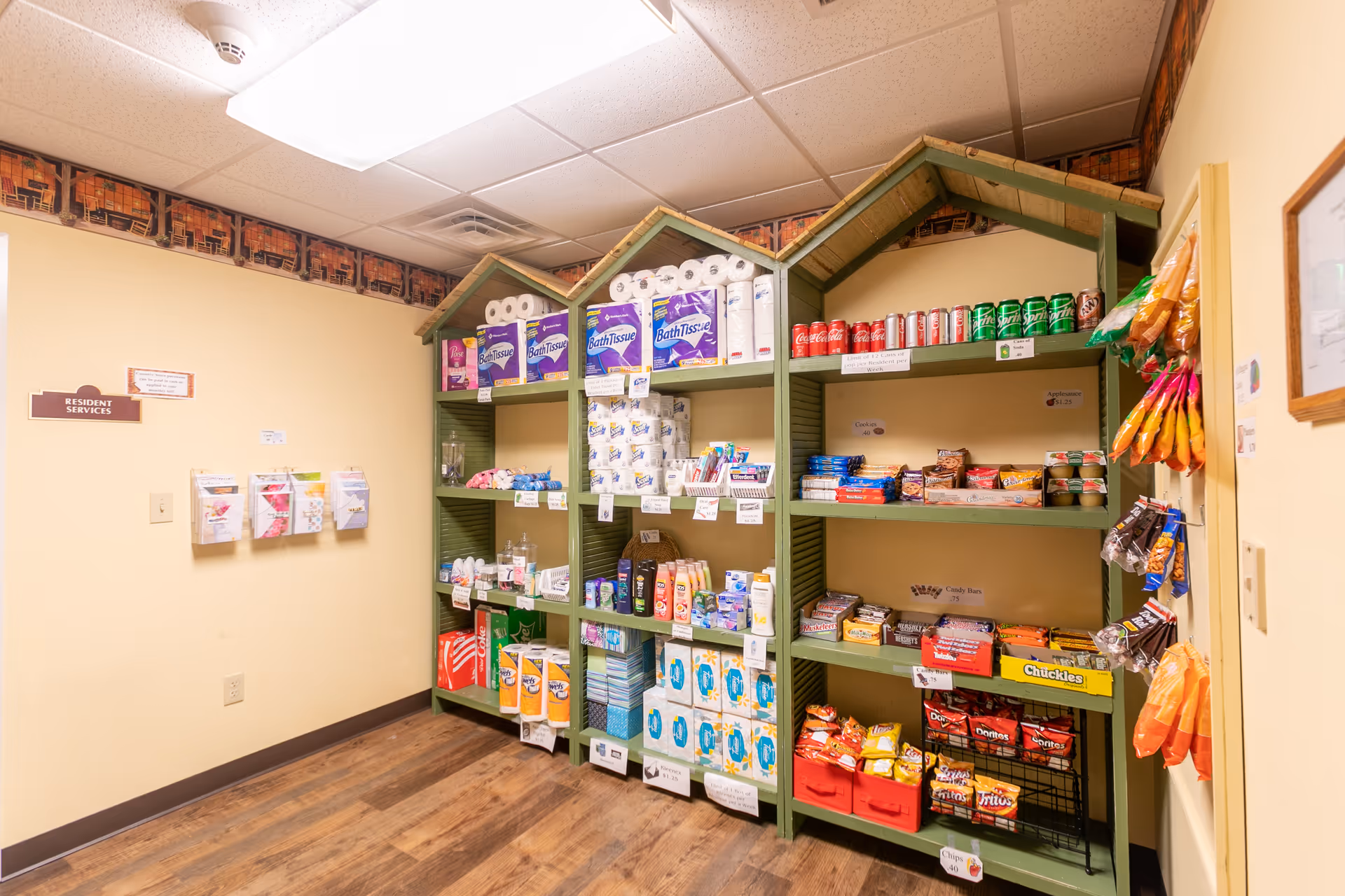 A small convenience area inside a senior living facility with shelves stocked with various items including paper towels, bath tissue, canned sodas, snacks, toiletries, and hygiene products. The shelves are green with a house-shaped top design. The walls are light yellow with a small sign labeled 'Resident Services' and some informational pamphlets mounted on the wall. The floor is wood-patterned.