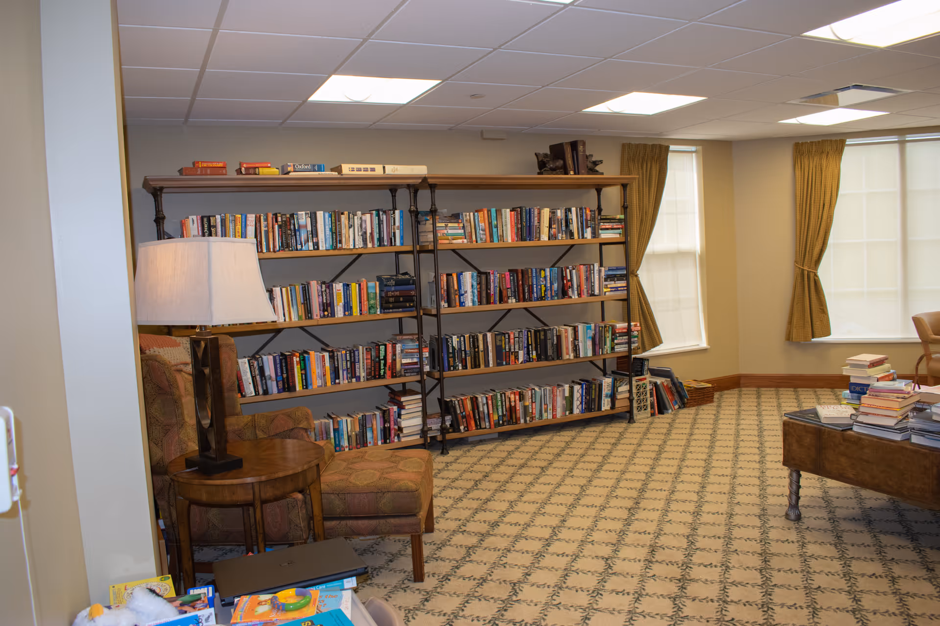 A cozy reading room with a large bookshelf filled with books against the wall, two windows with yellow curtains, a patterned carpet, a wooden table with stacks of books, and a comfortable armchair with a matching ottoman next to a table lamp.