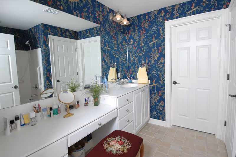A bathroom with blue floral wallpaper, a white vanity with a sink, a large mirror, and a countertop with various toiletries and a small plant. There is a cushioned bench with a floral pattern in front of the vanity. Two white doors are visible, one leading to a shower area.